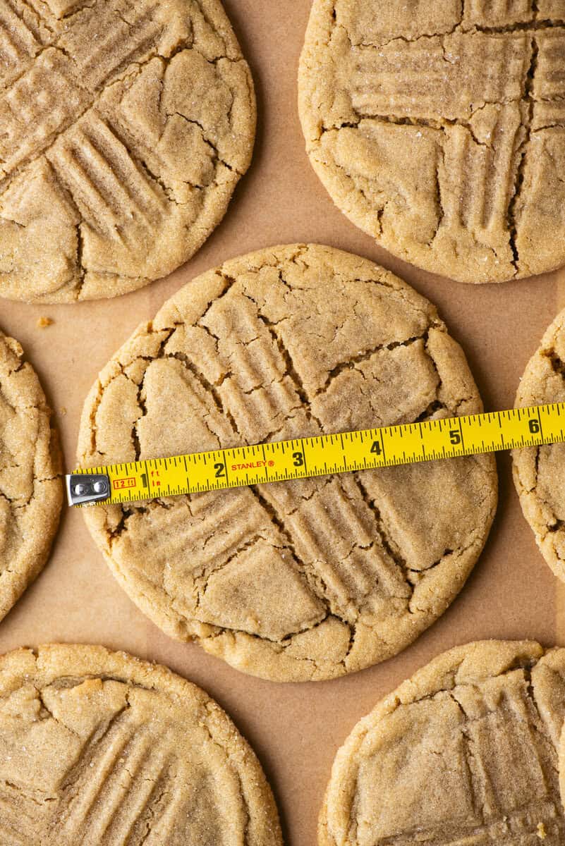 A yellow measuring tape lies across a large peanut butter cookie with a crisscross fork pattern, surrounded by similar cookies on brown parchment paper.