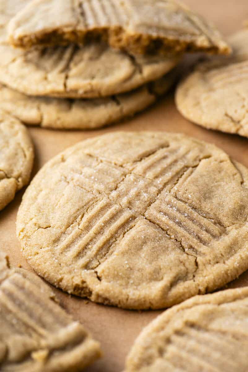Close-up of several peanut butter cookies with a crisscross fork pattern on top, arranged on parchment paper. One cookie is broken in half and stacked over another in the background.