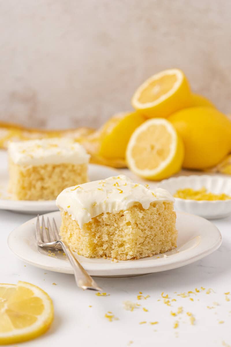 A square piece of lemon cake with white frosting and lemon zest sits on a white plate with a fork. In the background are more cake pieces and whole and sliced lemons.