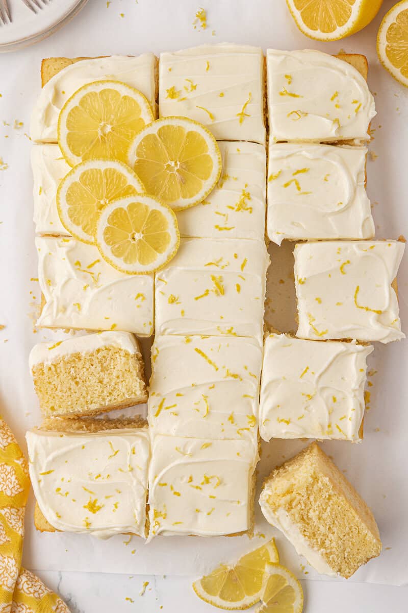 Slices of lemon cake with creamy white frosting are arranged on parchment paper. Some slices are topped with lemon zest and lemon slices, and a few pieces are separated from the main cake.