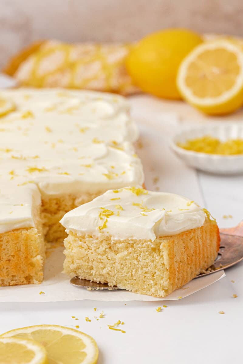 A frosted lemon sheet cake with a piece cut out and lifted, topped with lemon zest. Sliced lemons and grated zest are in the background. The cake has a moist, fluffy texture and creamy white icing.