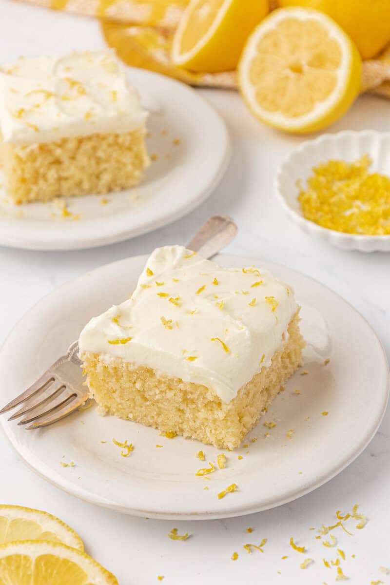 A slice of lemon cake with creamy white frosting and lemon zest sits on a white plate with a fork. More cake, lemon zest, and sliced lemons are in the background.