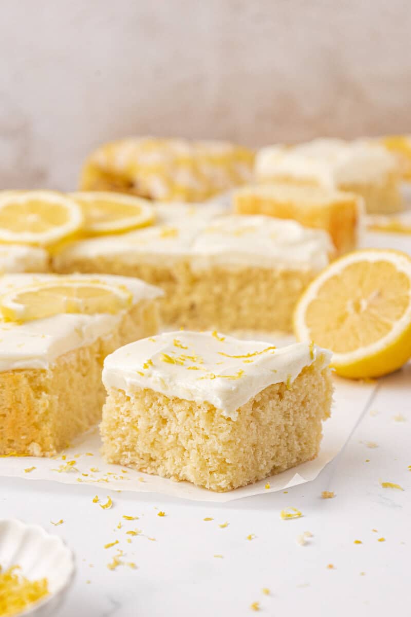 A close-up of lemon cake squares topped with white frosting and lemon zest, with fresh lemon slices and a halved lemon in the background on a white surface.