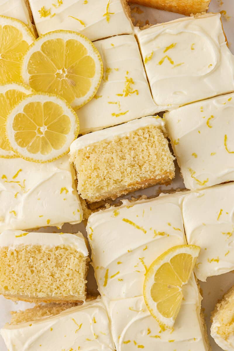 Lemon sheet cake slices with creamy white frosting, topped with lemon zest and garnished with fresh lemon slices, arranged closely together.