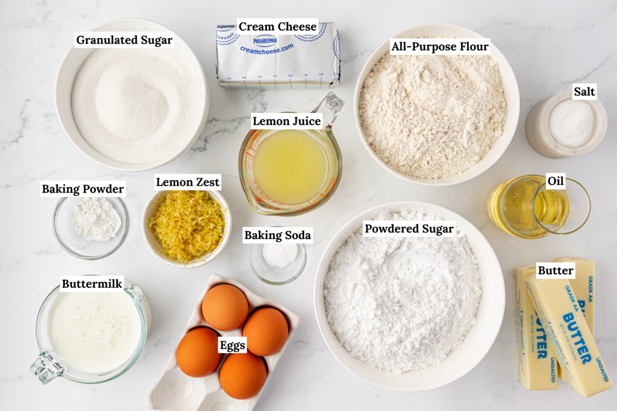 Top-down view of baking ingredients in bowls and containers on a white surface, including granulated sugar, cream cheese, flour, salt, oil, powdered sugar, butter, eggs, buttermilk, lemon juice, lemon zest, baking soda, and baking powder.