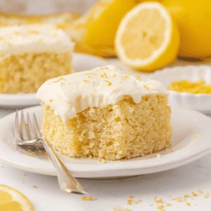 A square slice of moist lemon cake with creamy white frosting sits on a white plate with a fork. In the background are lemon slices and zest, adding a fresh, bright atmosphere.