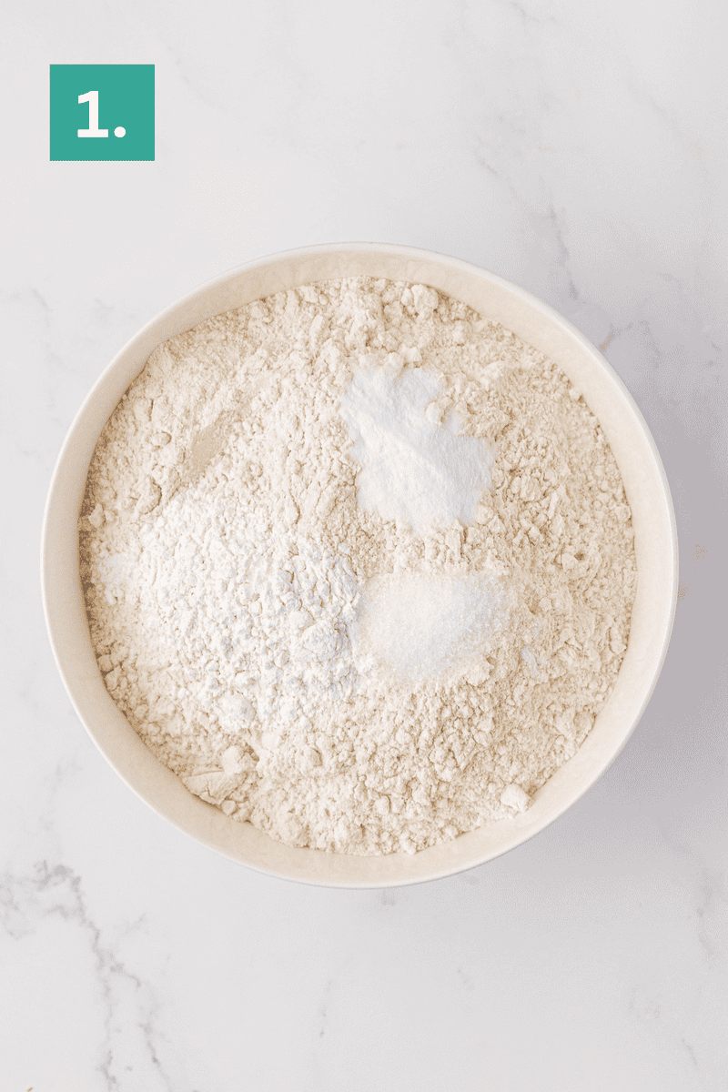 A white bowl filled with flour and white powdery ingredients sits on a marble surface. A green box with the number 1 is in the top left corner, indicating the first step in a process.