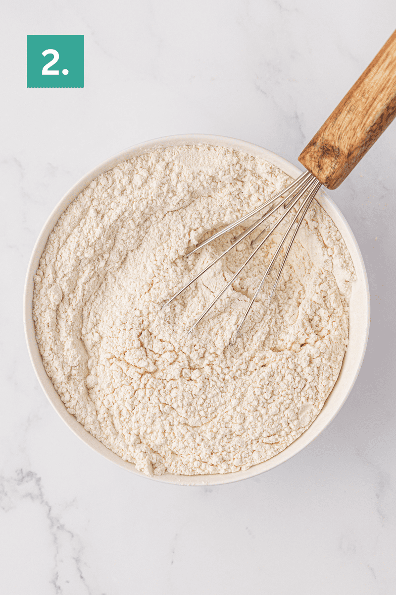 A bowl of flour mixture with a metal whisk and a wooden handle on a white surface. A green box with the number 2 is in the top left corner.