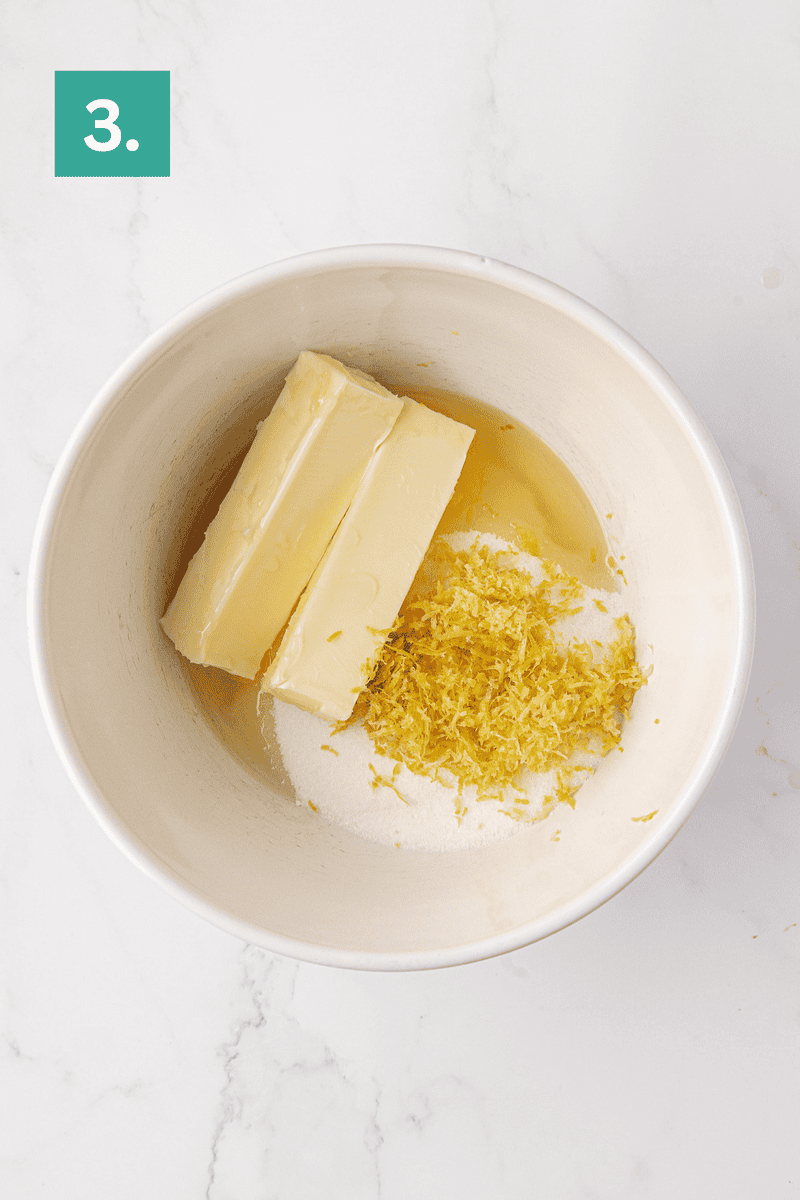 A mixing bowl with two sticks of butter, granulated sugar, lemon zest, and a liquid ingredient on a white surface. A green box in the top left corner displays the number 3.