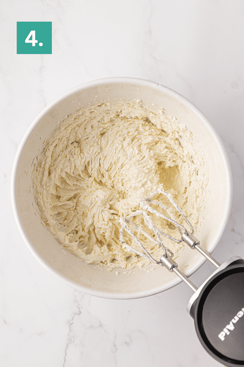A hand mixer with beaters sits in a white bowl of whipped, creamy mixture on a white surface. A green box labeled “4.” is in the top left corner.