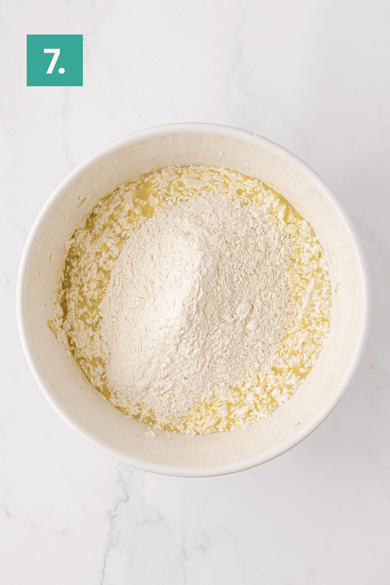 A mixing bowl containing wet ingredients with a mound of flour on top sits on a white surface. A green box labeled 7. appears in the top left corner, indicating this is step seven in a recipe.