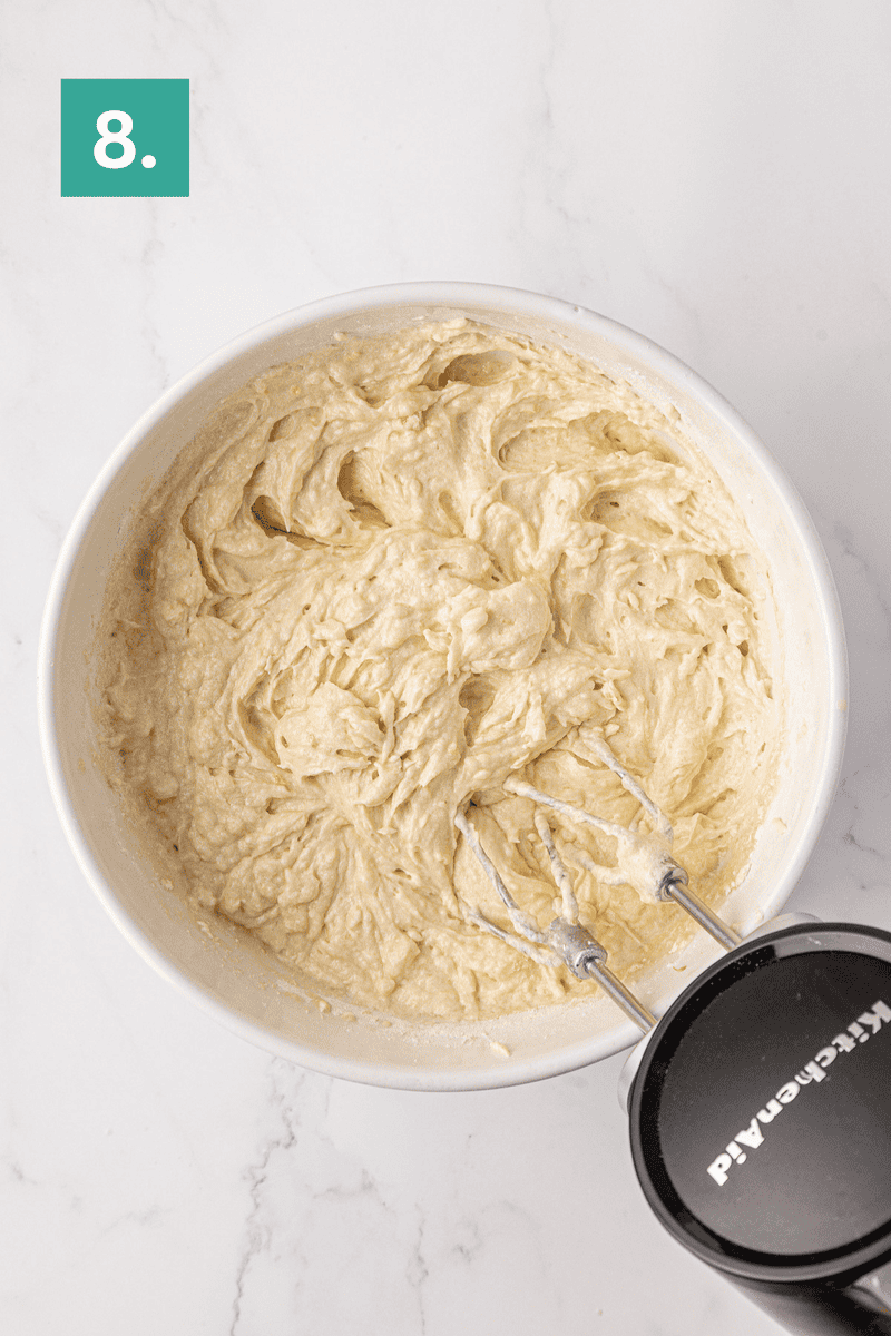 A mixing bowl filled with creamy cake batter, with a hand mixer’s beaters partially submerged. A green box with the number 8 is in the top left corner, indicating a recipe step.