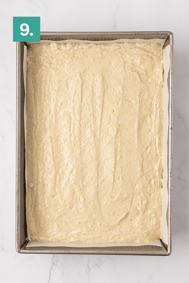 A rectangular baking pan lined with parchment paper is filled with an even layer of cake batter. A green square in the top left corner has the number 9 in white. The pan is on a white marble surface.