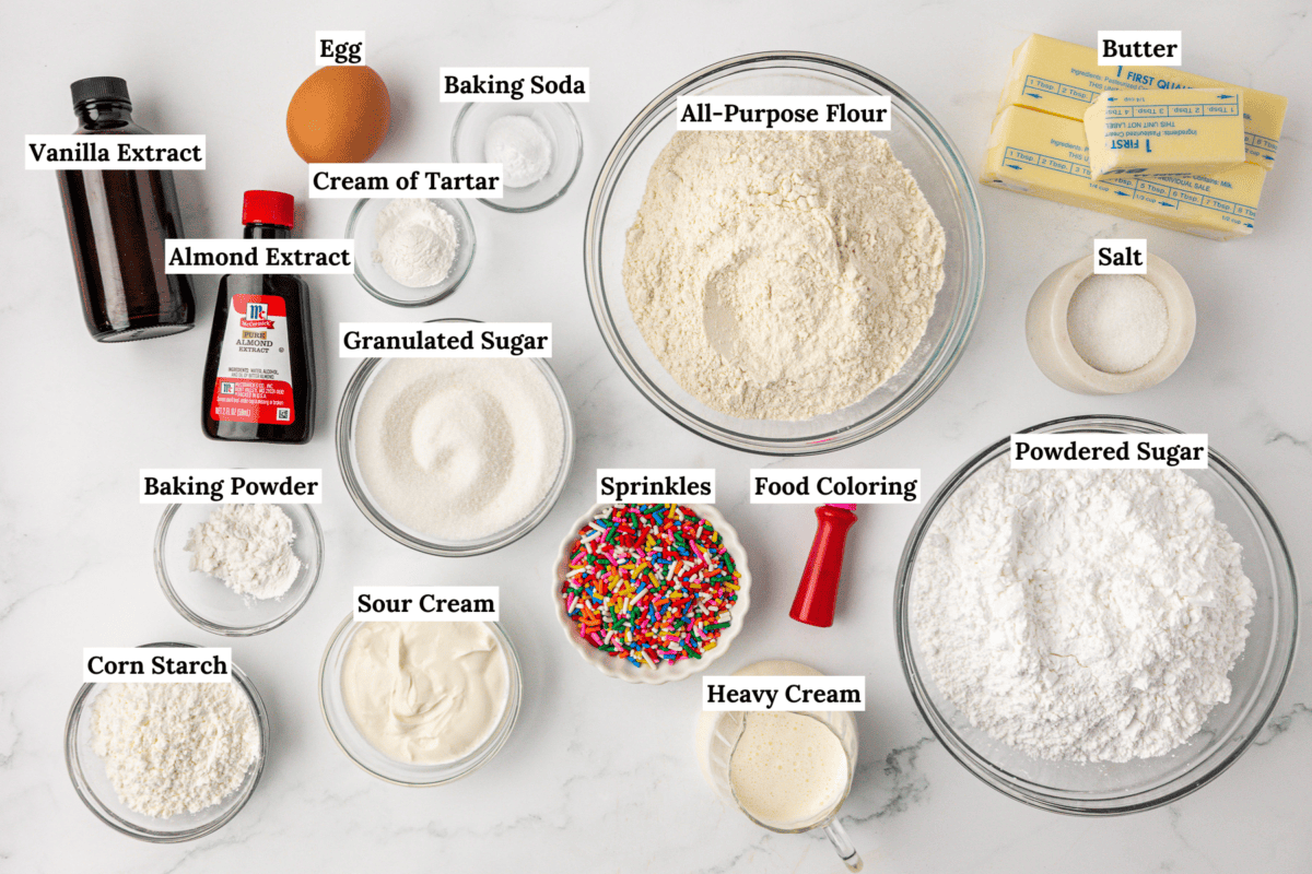 Top-down view of labeled baking ingredients in bowls and containers on a white surface, including flour, sugar, eggs, extracts, baking powder, baking soda, cream of tartar, sprinkles, butter, food coloring, sour cream, and heavy cream.