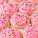 A batch of round sugar cookies topped with thick pink frosting and colorful rainbow sprinkles, arranged closely together.