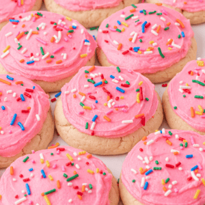 A batch of round sugar cookies topped with thick pink frosting and colorful rainbow sprinkles, arranged closely together.