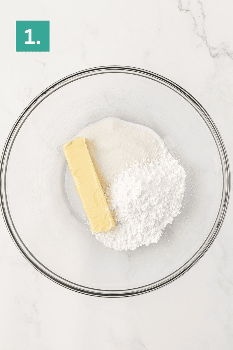 A clear glass mixing bowl containing a stick of butter, granulated sugar, and powdered sugar sits on a white marble surface. A green box labeled 1. is in the top left corner.