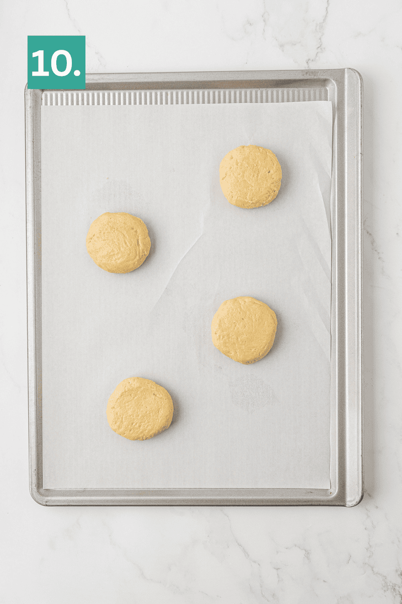 A baking sheet lined with parchment paper holds four evenly spaced, round raw dough discs. A green box in the top left corner displays the number 10. The background is a white marble surface.