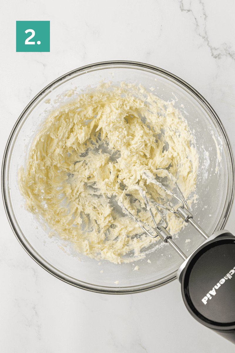 A mixing bowl with creamed butter and sugar being mixed by a handheld electric mixer on a white marble surface. A green box with the number 2 is in the top left corner.