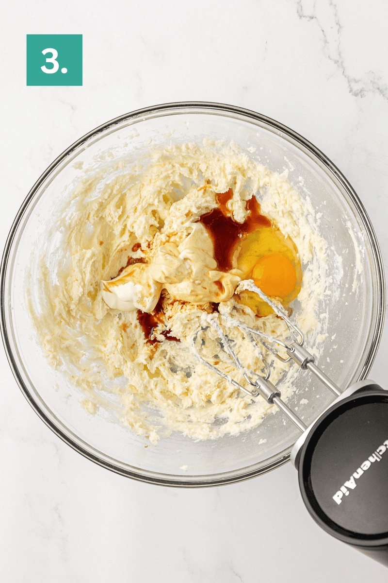 A glass bowl with mixed butter and sugar, an egg, vanilla, and sour cream being combined with a hand mixer. A green square with the number 3 is in the top left corner.