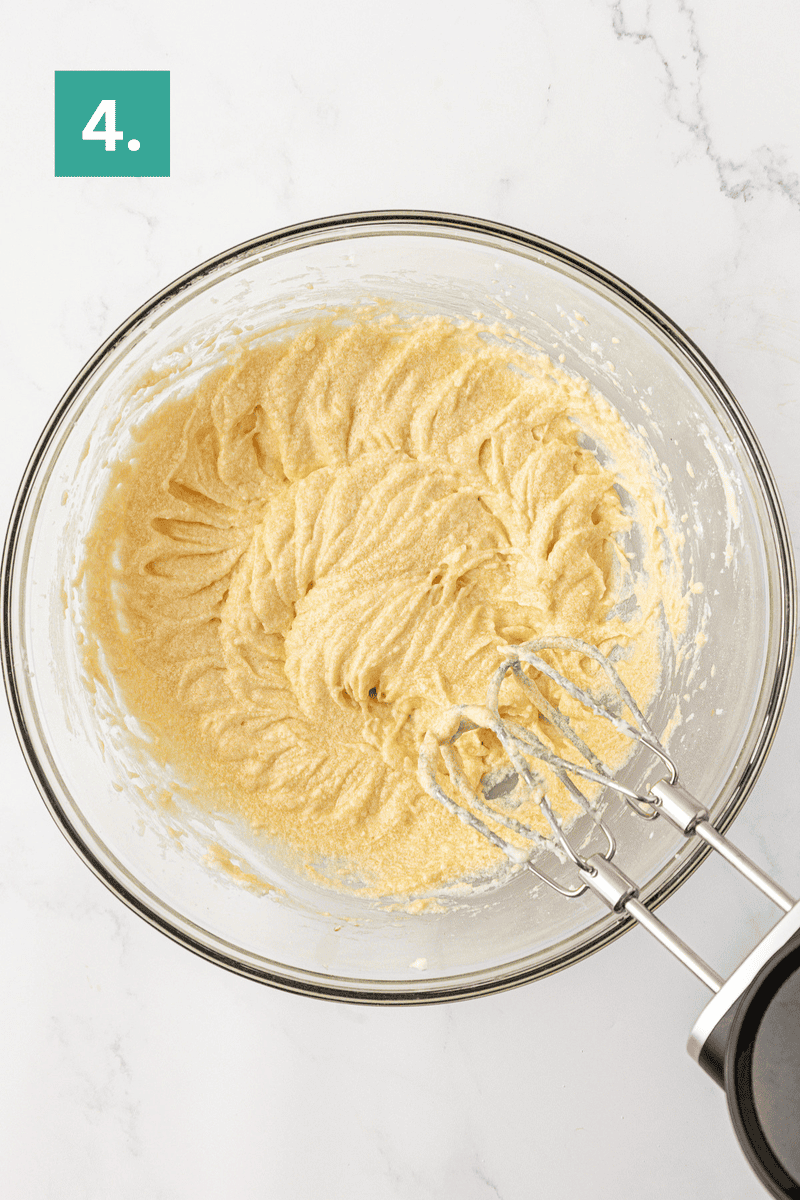 A glass bowl with creamy, whipped batter sits on a marble surface. An electric hand mixer with two beaters rests inside the bowl. A green box labeled 4. is in the top left corner.