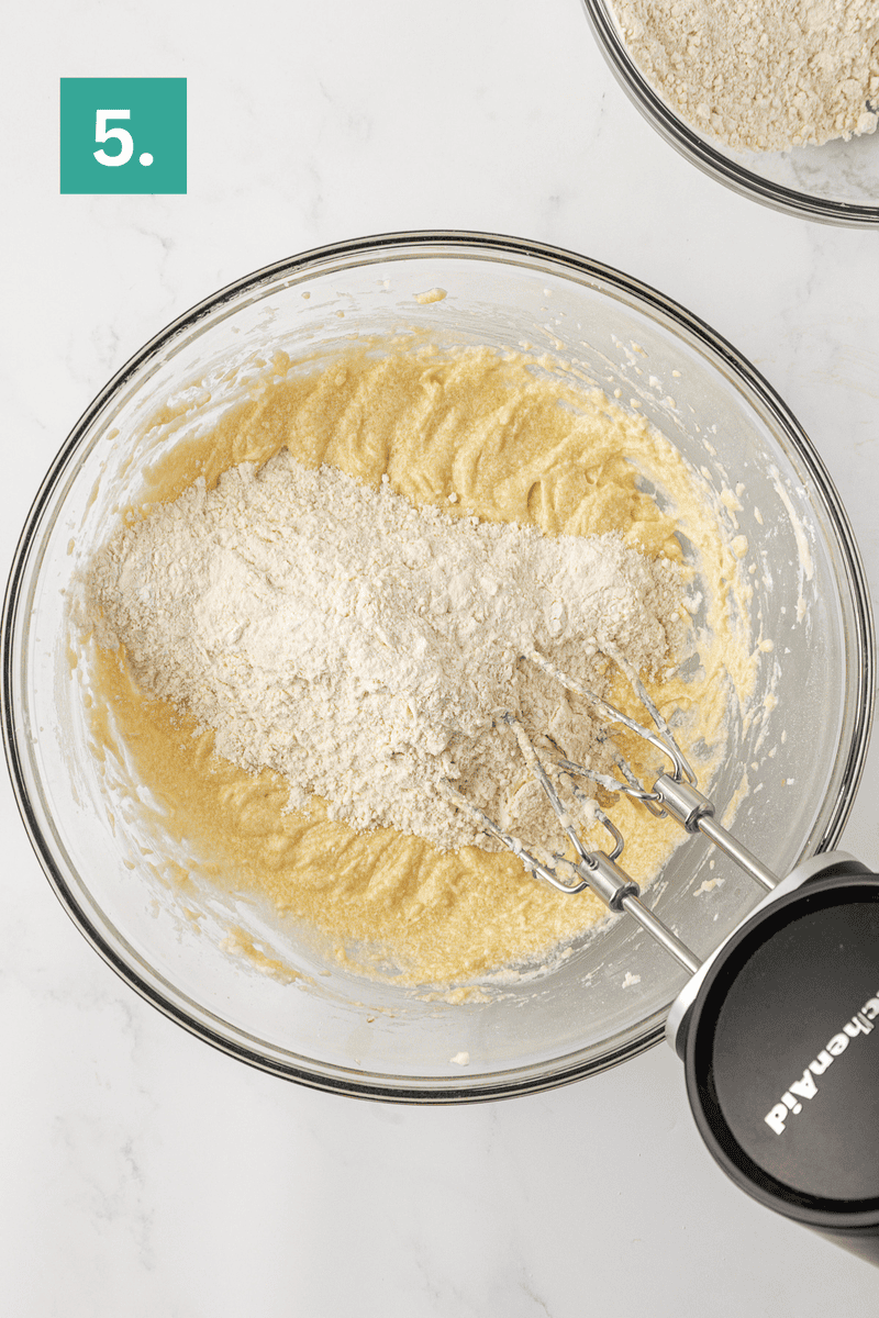A mixing bowl with creamy batter and flour being mixed with a hand mixer, viewed from above. A green square with the number 5 is in the top left corner. Another bowl with flour is partially visible at the top.