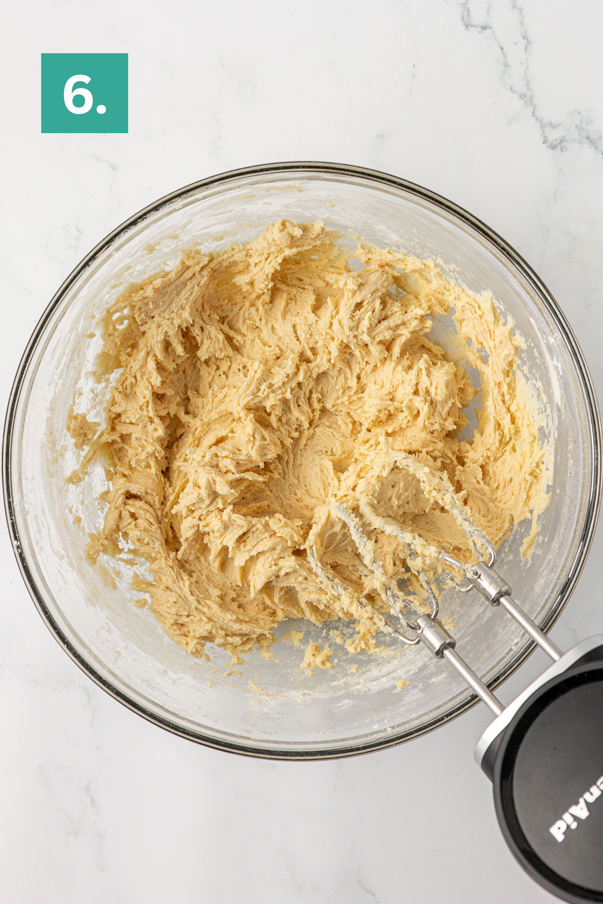 A glass bowl filled with creamy, mixed cookie dough sits on a marble surface. An electric hand mixer rests in the bowl. A green box with the number 6 is in the top left corner.