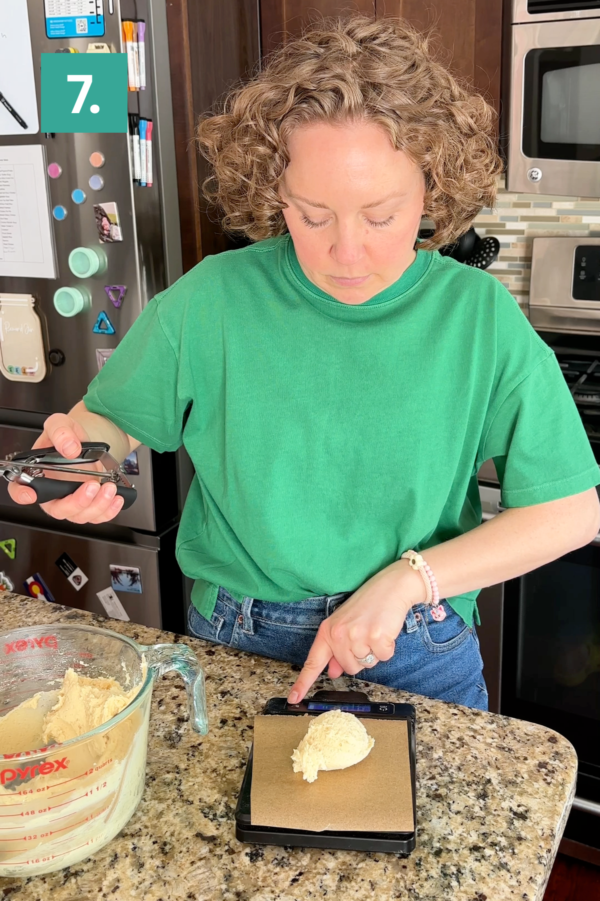 A person in a green shirt uses a kitchen scale to weigh a ball of dough on a counter, holding a phone in one hand. A glass measuring cup with more dough sits nearby. A number 7. appears in the top left corner.