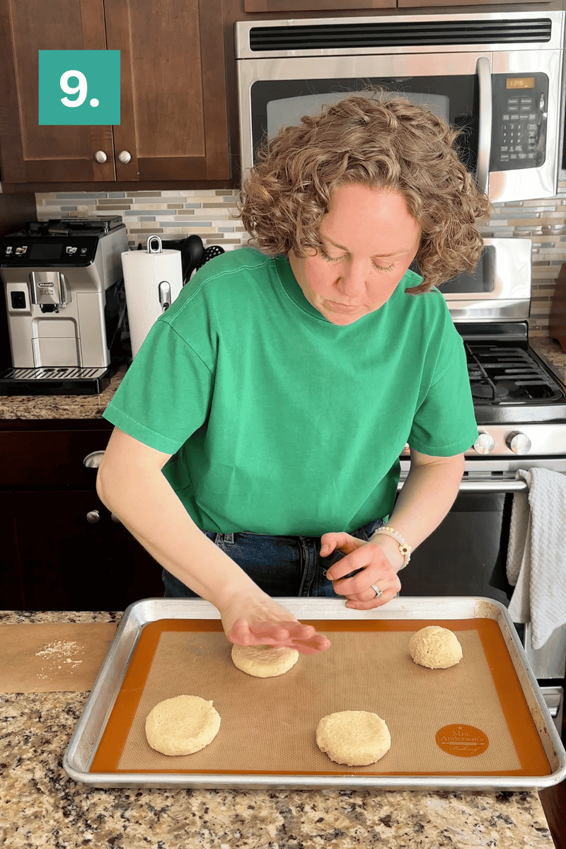 A person in a green shirt presses down on a round dough ball on a baking sheet, preparing biscuits in a kitchen. Three other dough rounds are also on the tray. A 9. label is in the top left corner.
