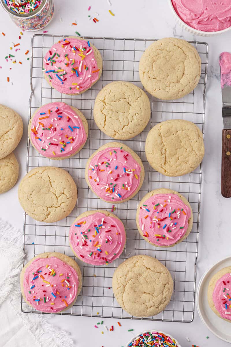 A cooling rack with sugar cookies, some plain and some topped with pink frosting and colorful sprinkles, on a white surface with scattered sprinkles and baking tools nearby.