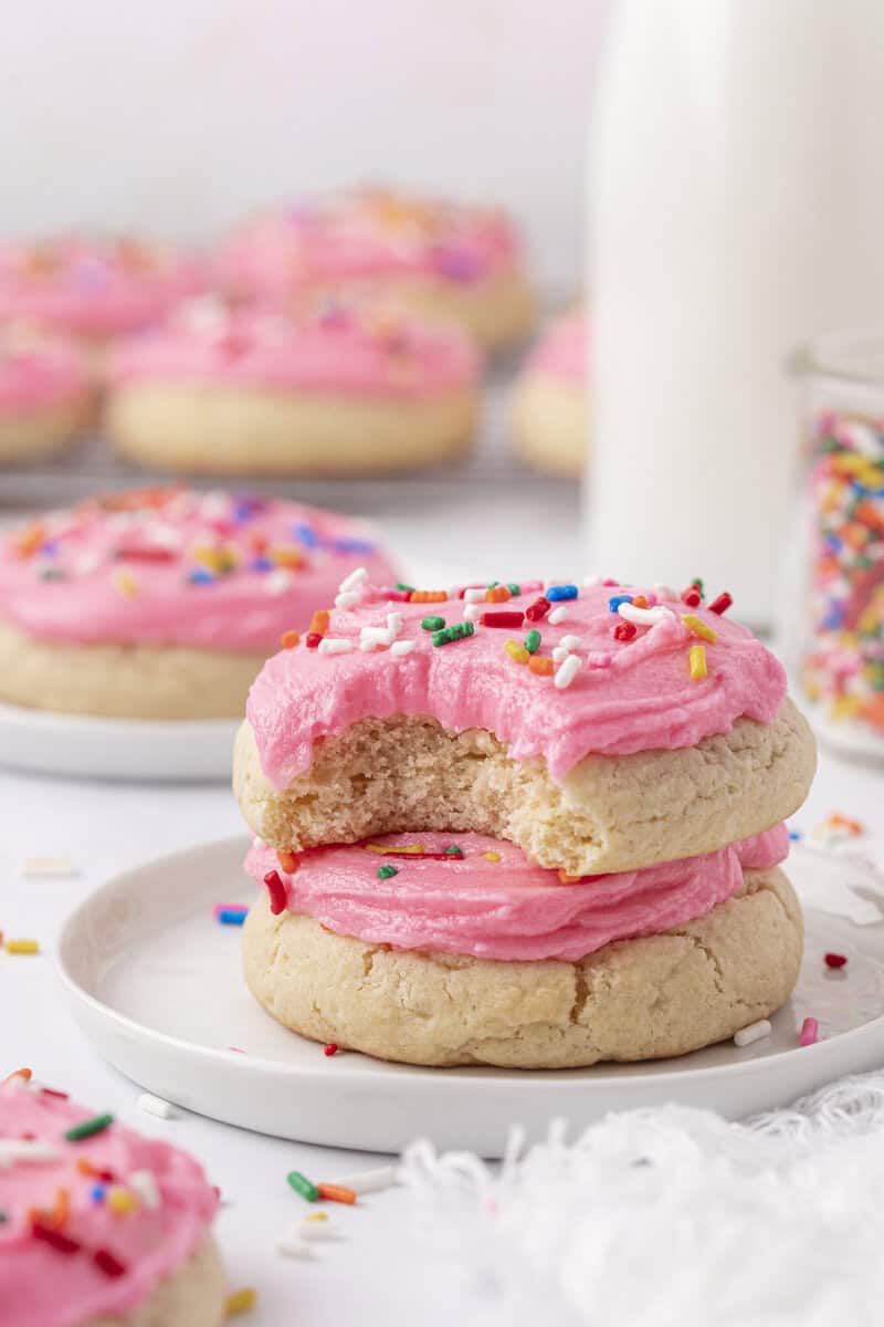 Two sugar cookies with pink frosting and colorful sprinkles are stacked on a white plate. The top cookie has a bite taken out of it. More frosted cookies and a glass of milk are visible in the background.