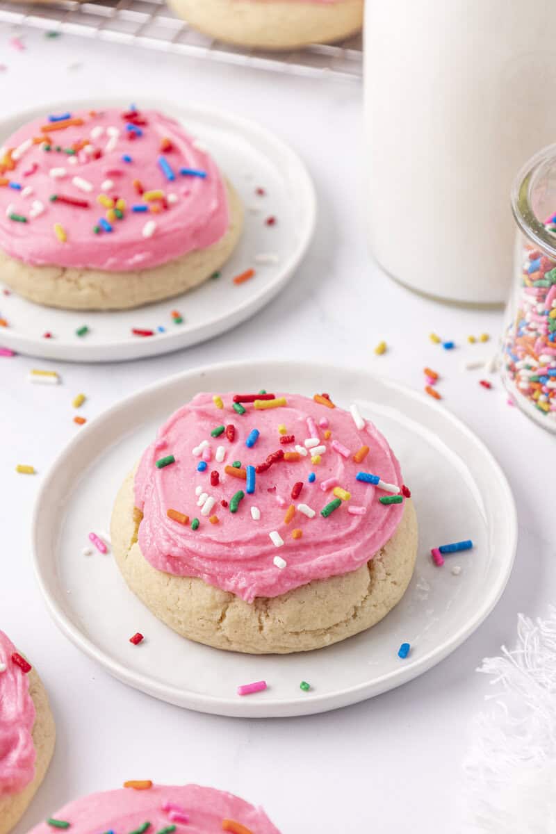 A plate with a sugar cookie topped with pink frosting and colorful sprinkles, next to a jar of more sprinkles, a glass of milk, and another frosted cookie in the background.