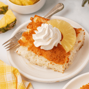 A slice of fluffy cake topped with a swirl of whipped cream and a pineapple ring sits on a white plate with a fork, next to a yellow checkered napkin.