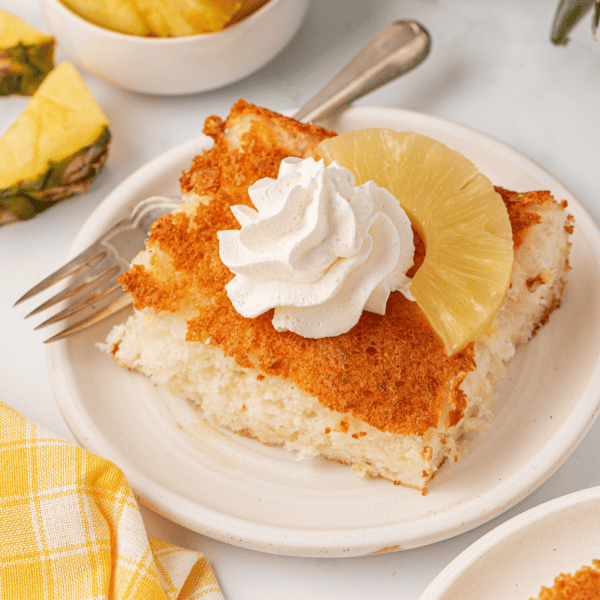 A slice of fluffy cake topped with a swirl of whipped cream and a pineapple ring sits on a white plate with a fork, next to a yellow checkered napkin.