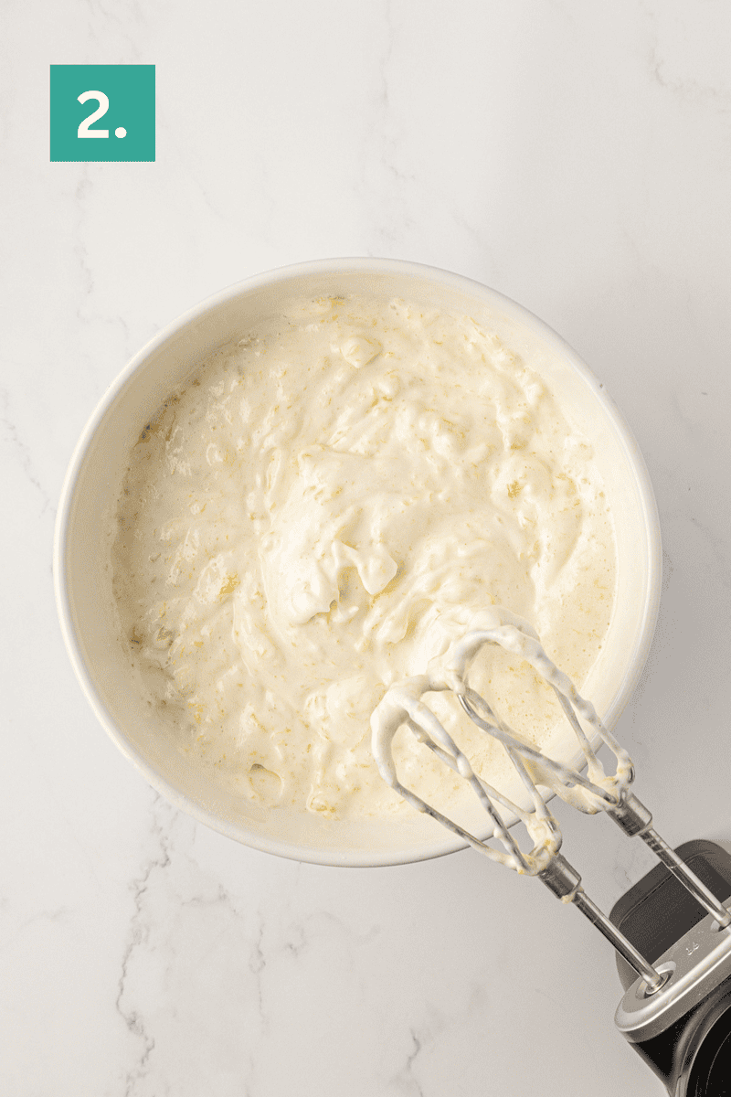 A white mixing bowl filled with creamy batter is being mixed with an electric hand mixer. A green square with the number 2 in white is in the top left corner. The background is a light marble surface.