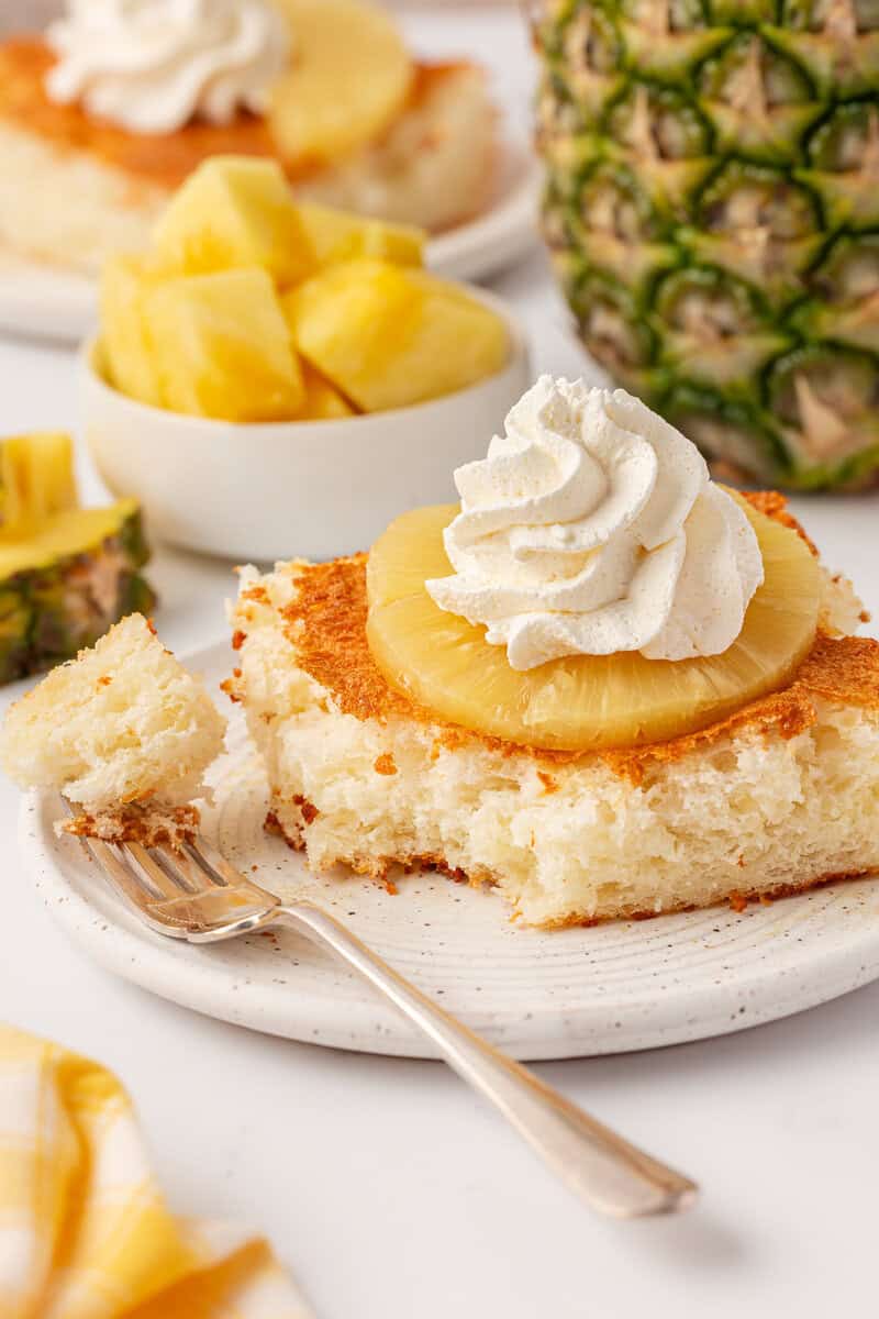 A slice of pineapple upside-down cake topped with a pineapple ring and whipped cream sits on a white plate beside a fork, with fresh pineapple pieces and a whole pineapple in the background.