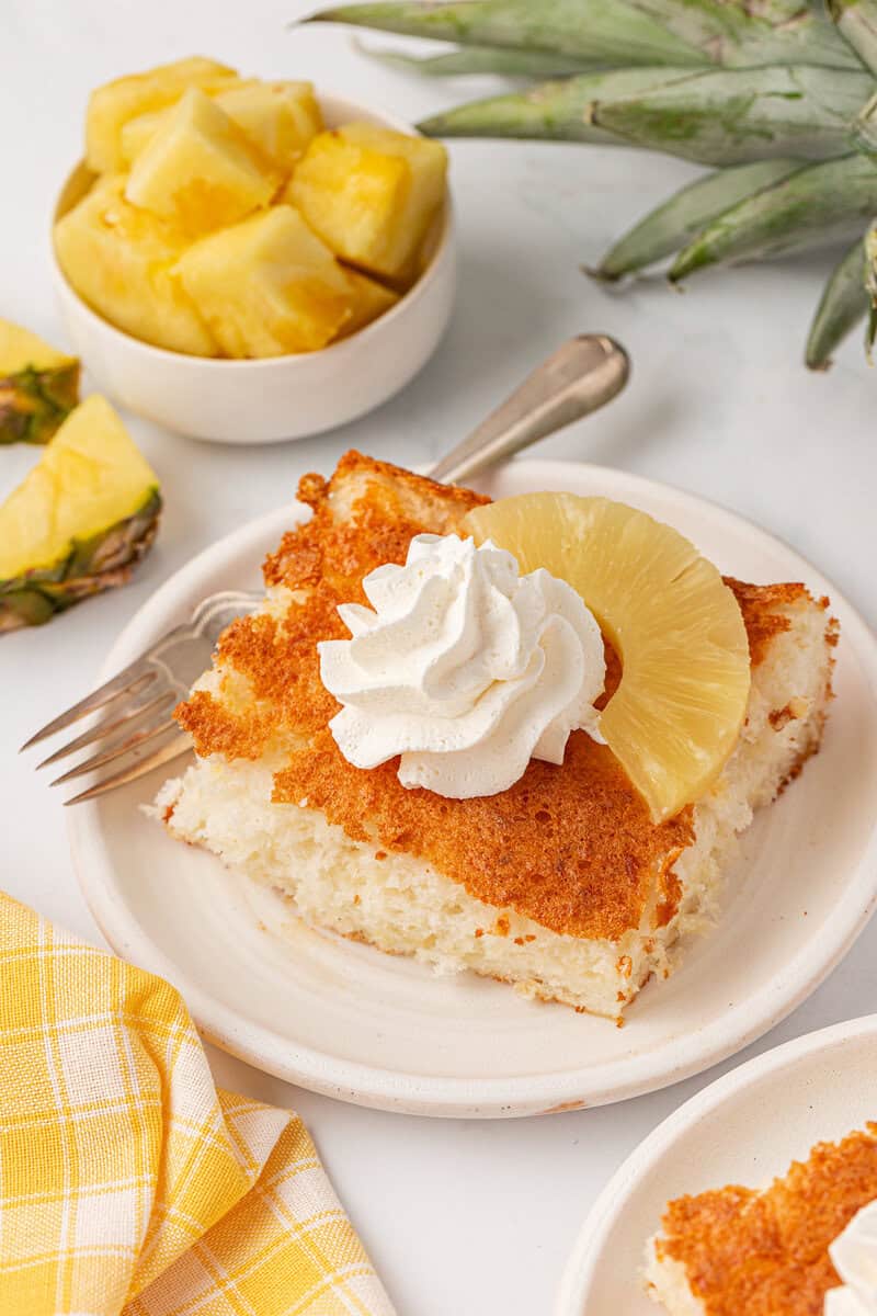 A slice of pineapple cake topped with whipped cream and a pineapple ring on a plate, with a fork, a bowl of pineapple chunks, and a yellow checkered napkin nearby.
