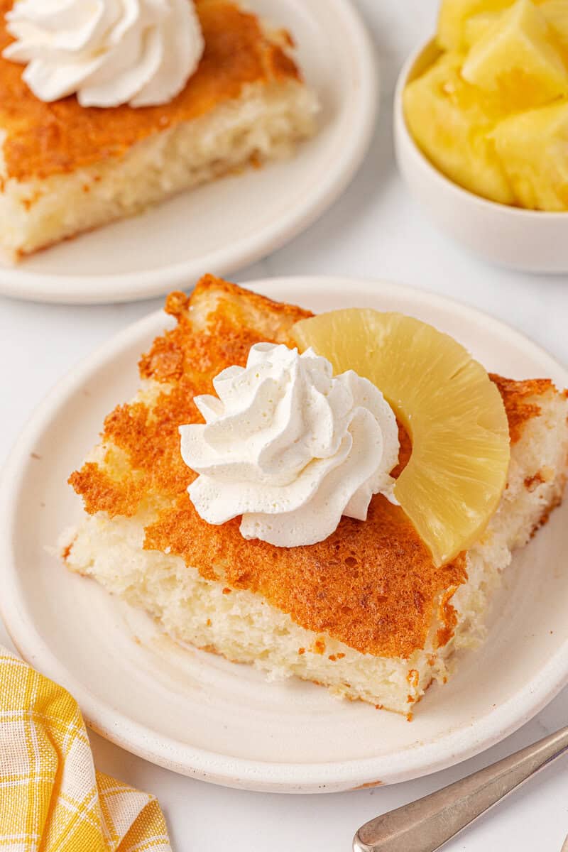 A slice of golden pineapple cake topped with a swirl of whipped cream and a pineapple ring sits on a white plate, with a bowl of pineapple chunks and another cake slice in the background.