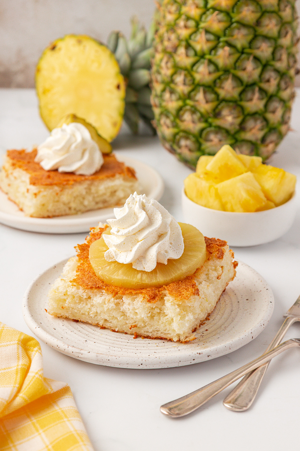 A slice of pineapple upside-down cake topped with pineapple and whipped cream sits on a plate. In the background are another cake slice, a whole pineapple, pineapple chunks in a bowl, and a yellow checkered napkin.
