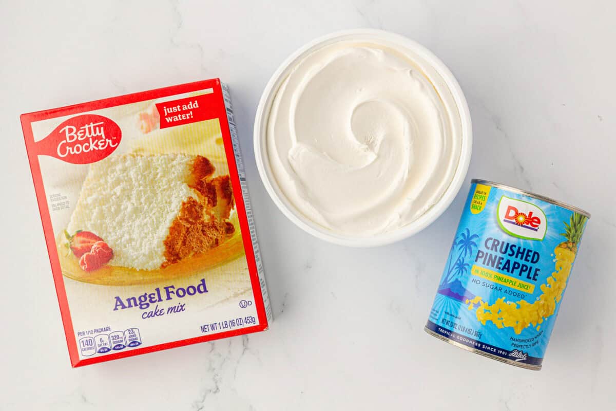 A box of Betty Crocker Angel Food cake mix, a tub of white whipped topping, and a can of Dole crushed pineapple are arranged on a white surface.