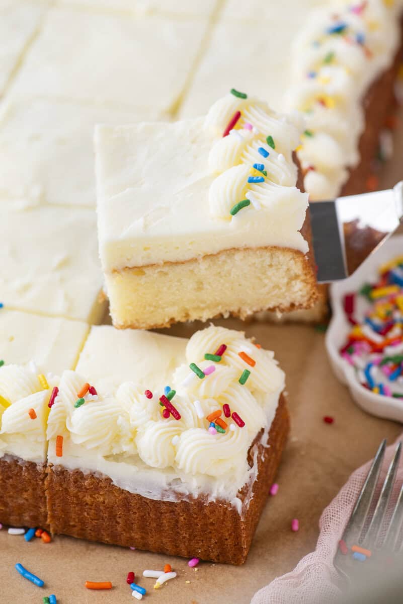 A square slice of frosted vanilla cake with piped white icing and colorful sprinkles is being lifted from a larger cake, with more sprinkles scattered nearby.