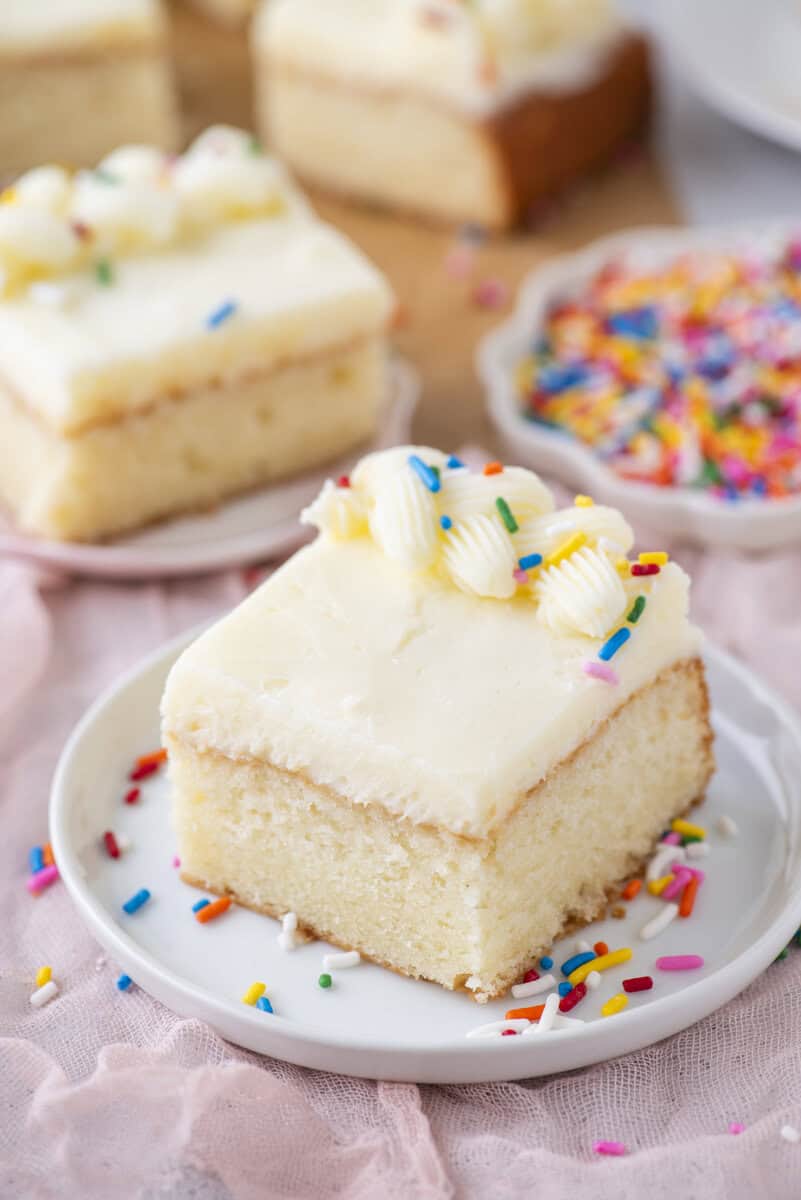A square piece of vanilla cake with white frosting and colorful sprinkles sits on a white plate. More slices of cake and a small bowl of sprinkles are in the background.