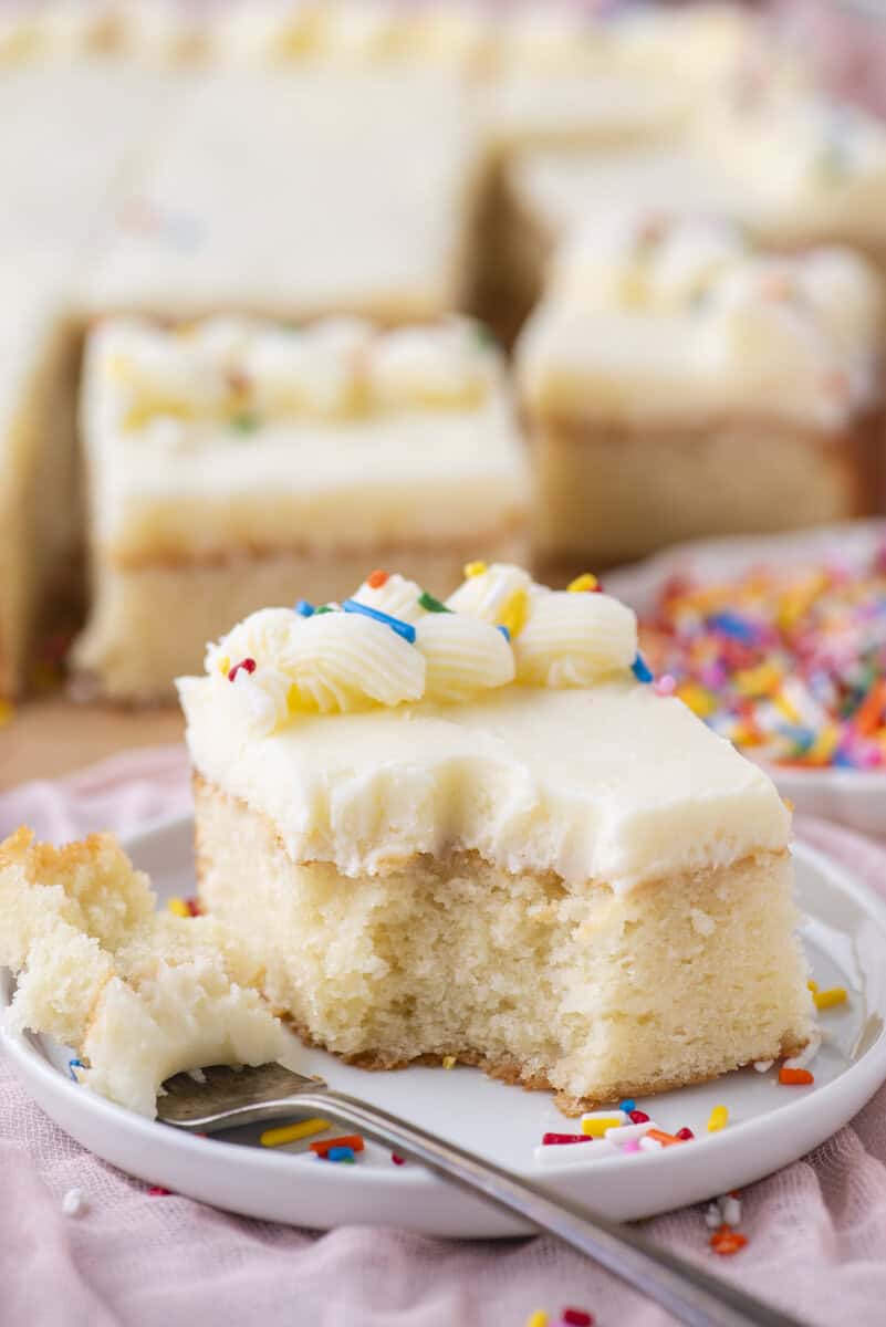 A slice of vanilla cake with white frosting and colorful sprinkles sits on a white plate, with a bite taken out and a fork nearby. More cake pieces and sprinkles are blurred in the background.