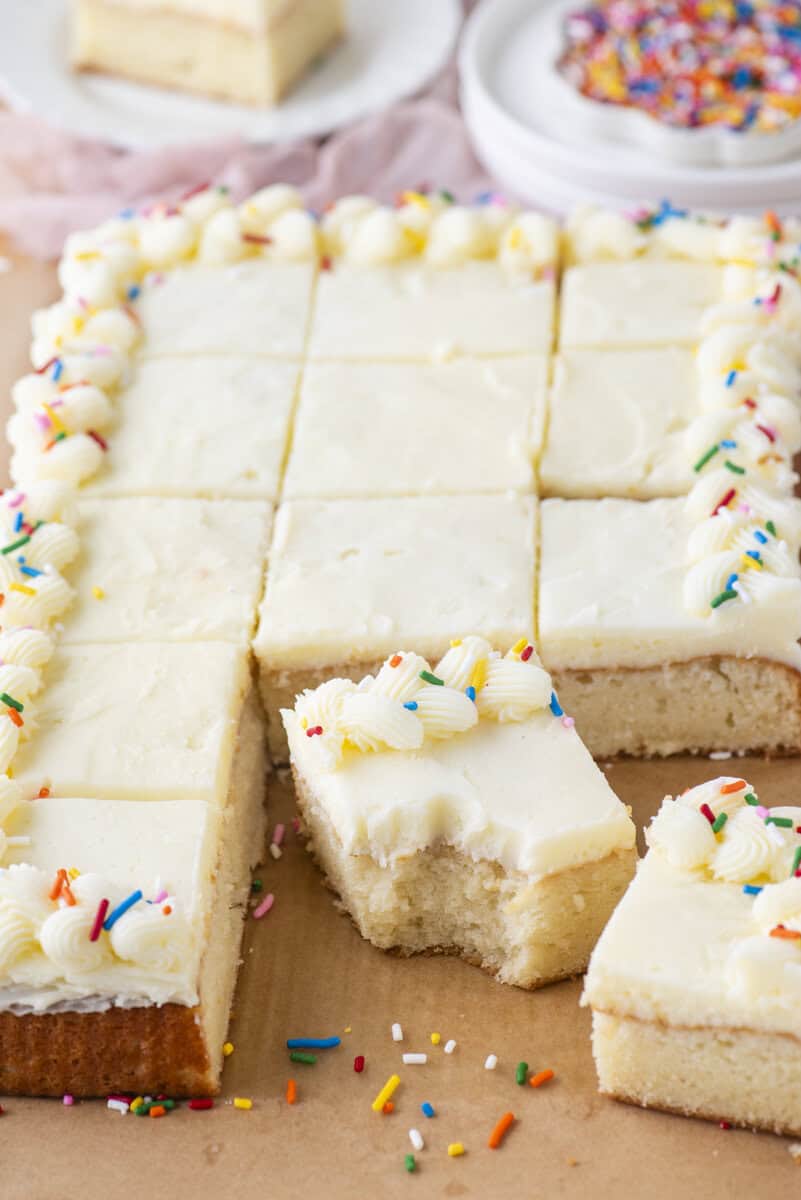 A rectangular white frosted sheet cake, cut into squares, with piped frosting and colorful sprinkles on the edges. One piece is pulled out in front with a bite taken from it. Plates with sprinkles are in the background.