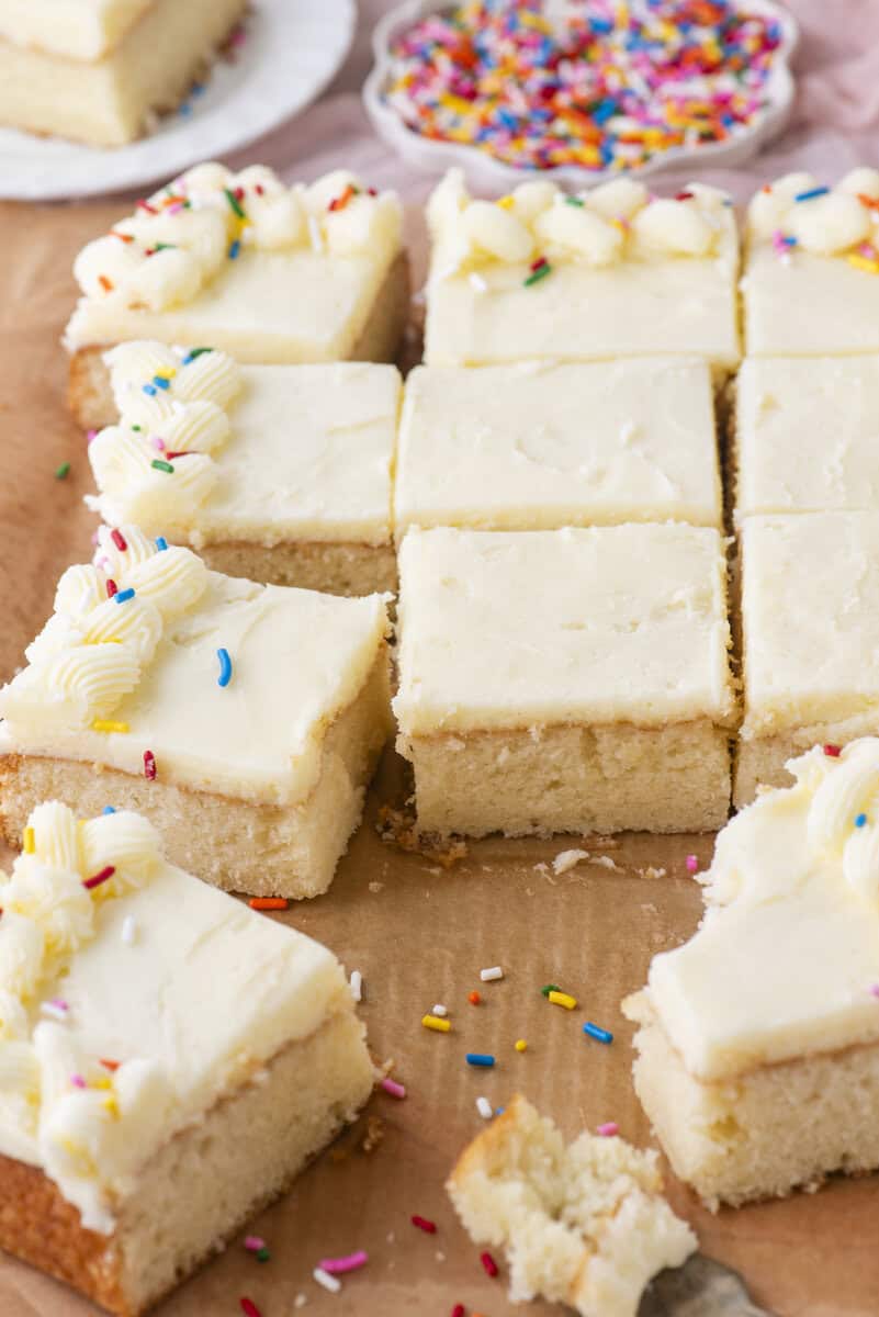 Nine thick, square pieces of vanilla cake with white frosting, some topped with colorful sprinkles and piped frosting. A bite is missing from one piece. Plates with sprinkles are visible in the background.