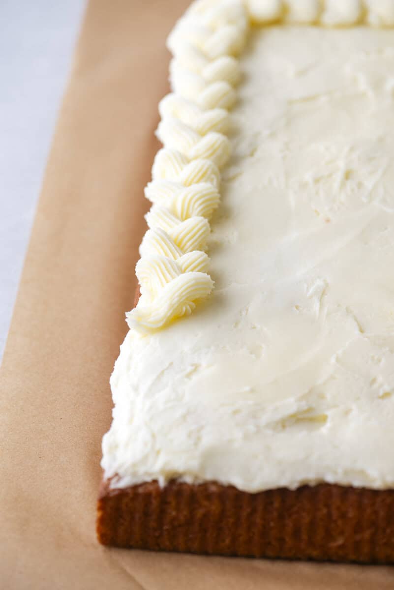 A close-up of a rectangular cake with smooth white frosting and a decorative piped border of icing along the edge, displayed on a sheet of parchment paper.