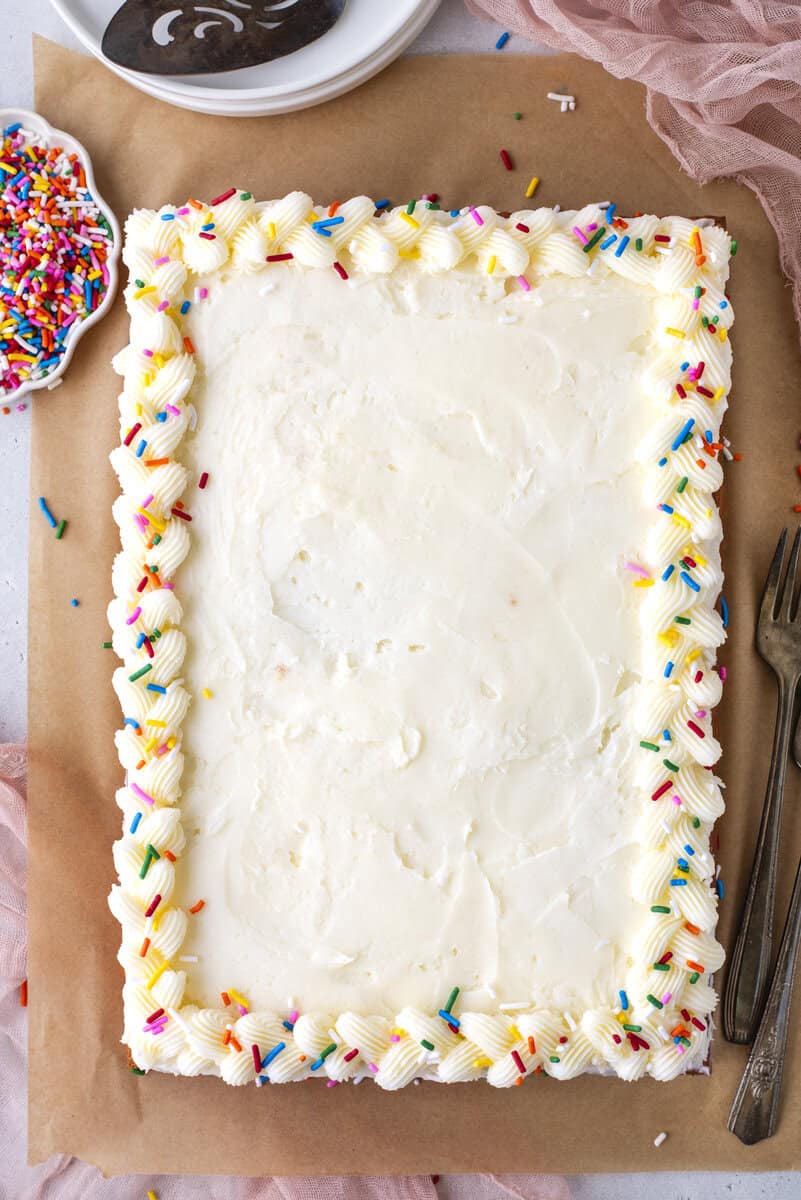 A rectangular sheet cake with white frosting, decorated with a piped border and colorful rainbow sprinkles, sits on brown parchment paper beside a small dish of extra sprinkles, a cake server, plates, and forks.