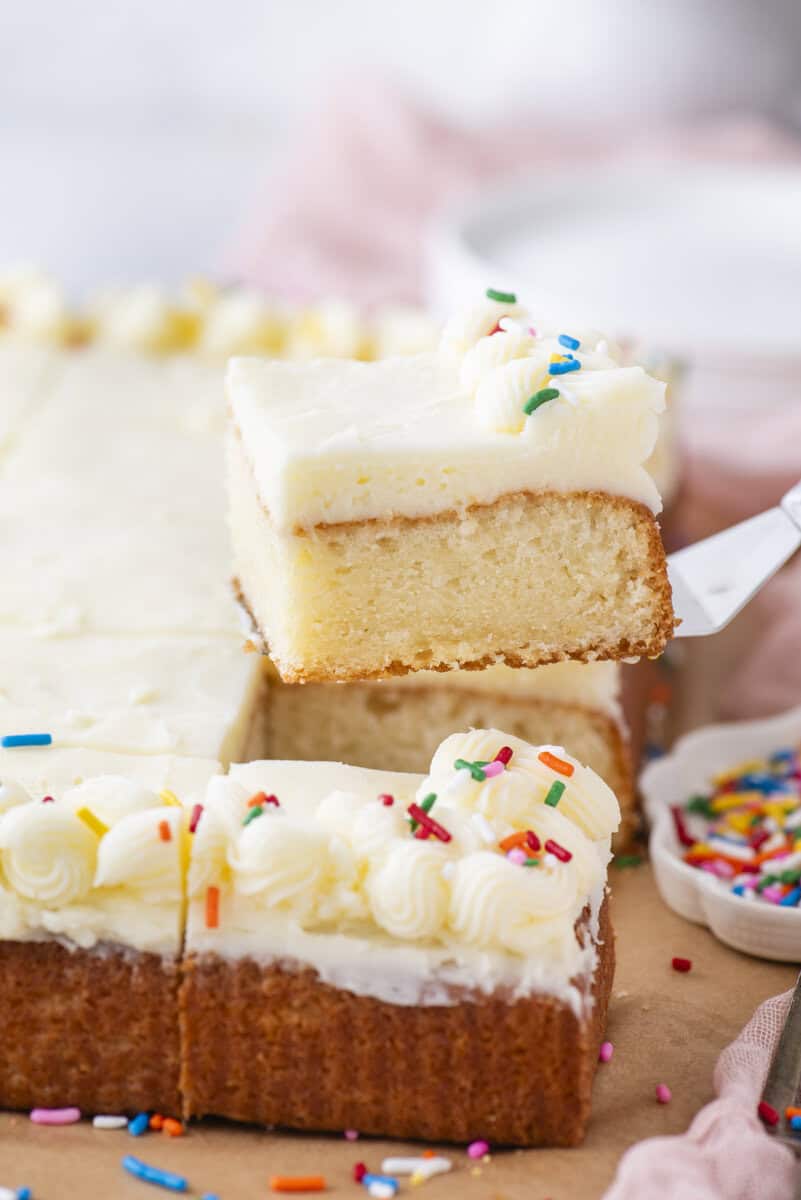 A slice of frosted vanilla sheet cake with colorful sprinkles is being lifted from the cake, which is decorated with piped white frosting and more sprinkles. A small dish of sprinkles sits nearby.
