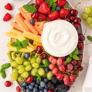 A platter of assorted fresh fruits including strawberries, cherries, grapes, kiwi, blackberries, blueberries, cantaloupe, and honeydew, arranged around a bowl of creamy dip, garnished with mint leaves.