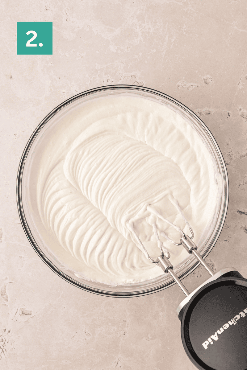 A glass bowl filled with whipped cream sits on a light countertop. An electric hand mixer with beaters, still attached, rests in the bowl, showing creamy, smooth peaks. A green square with the number 2 is in the corner.